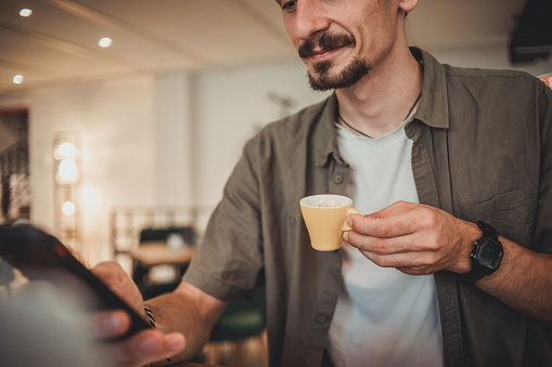 A young hipster man in a cafe drinking coffee and using a mobile phone to surf the Internet or correspond on social networks