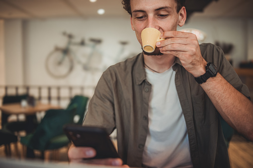 A young hipster man in a cafe drinking coffee and using a mobile phone to surf the Internet or correspond on social networks