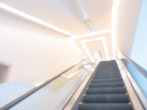 Blurred photo scene of empty escalator going up to the upstair with ceiling lighting decoration. Abstract blur background with escalator in white clean modern hall building.