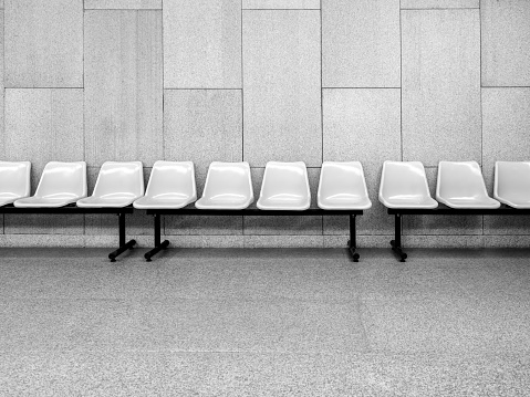 Row of empty white waiting plastic chairs seats with black iron legs seats on grey tiles and concrete wall background inside the modern building with nobody.