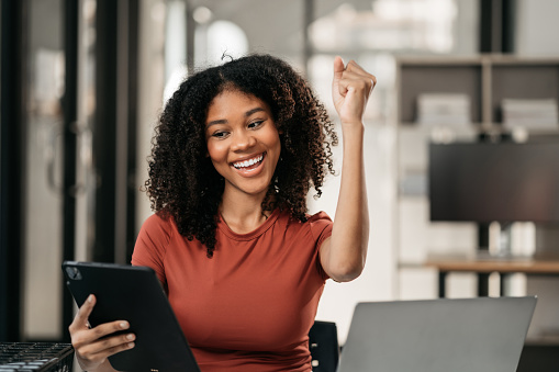 Excited african, american woman sitting at table feeling happy black woman overjoyed accepting mail at laptop promoted at home surprised girl reading good news.