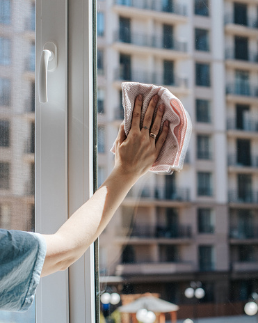 a woman's hand wipes a window with a napkin, another house is visible behind it, a sunny day