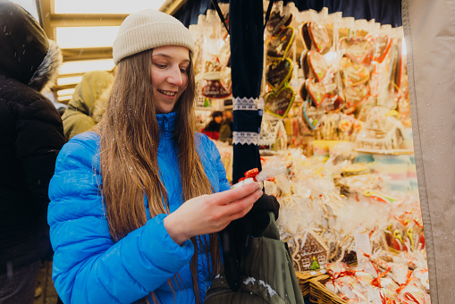 Portrait of female with long hair in hat and blue down jacket contemplating Christmas market in Krakow city, choosing souvenirs to buy during snowy winter weather