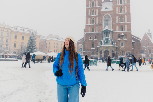 Front view of smiling female with long hair in hat and blue jacket walking an old streets of Krakow city, admiring snowfall and magical Christmas time