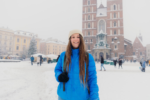 Front view of smiling female with long hair in hat and blue jacket walking an old streets of Krakow city, admiring snowfall and magical Christmas time