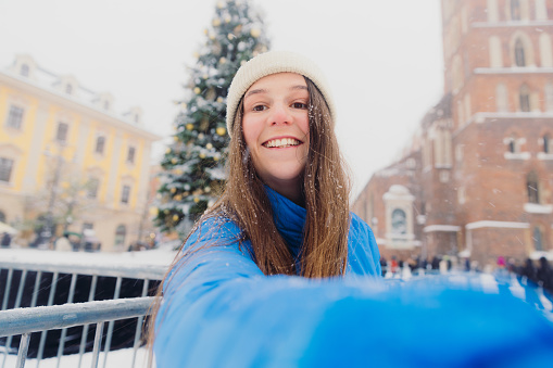 Self-portrait of smiling female with long hair in hat and blue jacket walking by Christmas Tree at old streets of Krakow city, admiring snowfall and magical winter time
