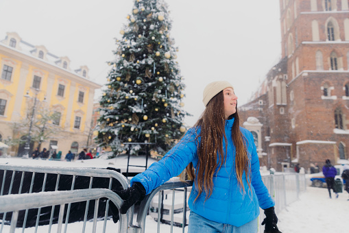 Low-angle view of smiling female with long hair in hat and blue jacket walking by Christmas Tree at old streets of Krakow city, admiring snowfall and magical winter time