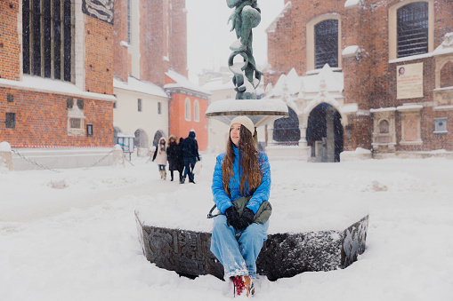 Front view of smiling female with long hair in hat and blue jacket sitting on fountain at old streets of Krakow city, admiring snowfall and magical winter time