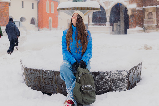 Front view of smiling female with long hair in hat and blue jacket sitting on fountain at old streets of Krakow city, admiring snowfall and magical winter time