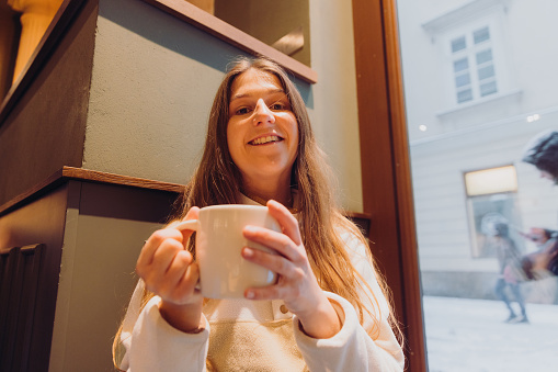 Low-angle view of female with long hair holding cup with coffee with window view of snow falling the old streets