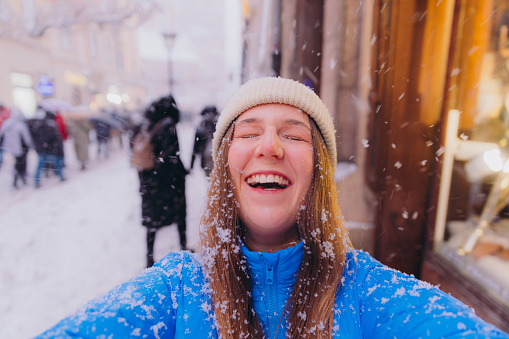 Self-portrait of smiling female with long hair in hat and blue jacket walking at old streets of Krakow city making video of herself, admiring snowfall and magical winter time