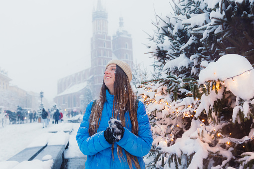 Front view of smiling female with long hair in hat and blue jacket walking an old streets of Krakow city, admiring snowfall and magical winter time with Christmas decoration around