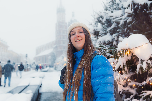 Side view of smiling female with long hair in hat and blue jacket walking an old streets of Krakow city, admiring snowfall and magical winter time with Christmas decoration around