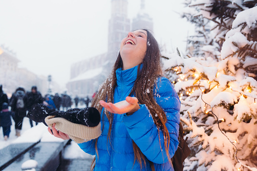 Portrait of smiling female with long hair in hat and blue jacket walking an old streets of Krakow city catching snowflakes by tongue, admiring snowfall and magical winter time with Christmas decoration around