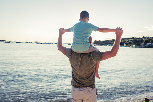 Cute little caucasian boy from behind cheering with arms outstretched while sitting on his father's shoulders at the beach. Playful young child from the back having fun and bonding with dad outdoors. The innocence of childhood.