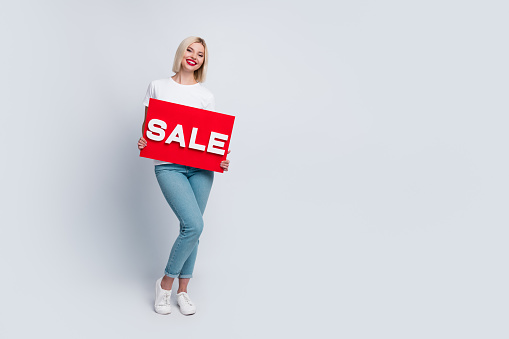 Full size photo of optimistic satisfied woman dressed white t-shirt holding sale red board in arms isolated on white color background.
