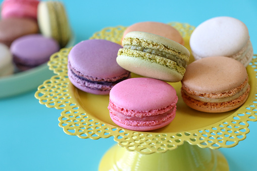 Stock photo showing close-up view of multi coloured macarons displayed on a yellow cake stand and a pastel blue plate against a blue background. Brown chocolate, white vanilla, purple blueberry, green pistachio, orange, pink strawberry flavoured meringues.