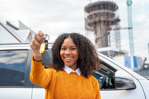 Black women with short, curly hair rent a car from the airport to travel by driving by herself. Rental or buying car concept. Holding keys to her new car.
