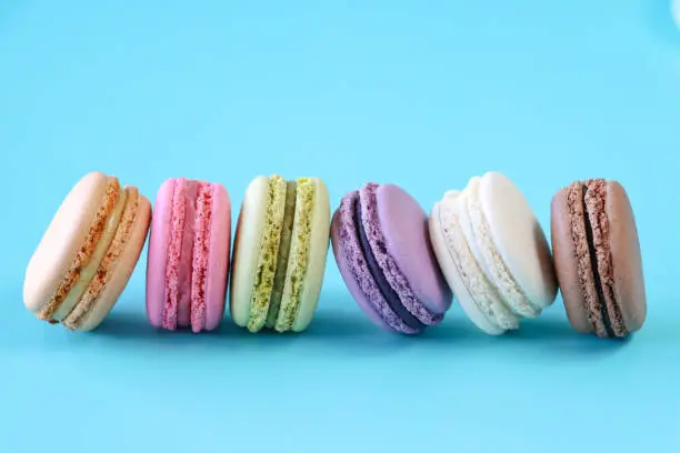 Photo of Image of unsteady row of six macarons on edge, multi coloured macarons filled with flavoured butter cream, chocolate, vanilla, blueberry, pistachio, orange, strawberry flavoured meringues, blue background, elevated view, copy space