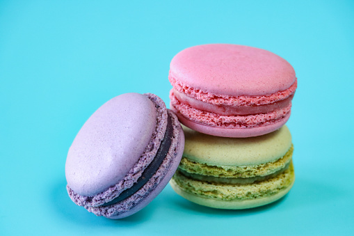 Stock photo showing close-up view of three multi coloured macarons on a blue background. Green pistachio, purple blueberry and pink strawberry flavoured meringues.