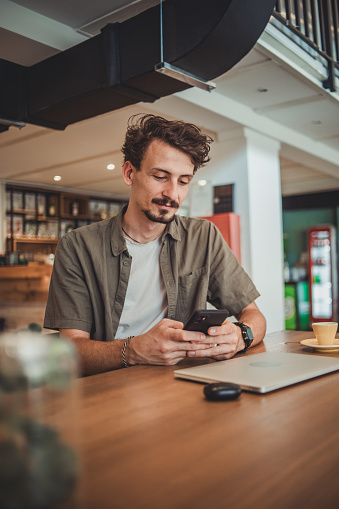 A young hipster man in a cafe drinking coffee and using a mobile phone to surf the Internet or correspond on social networks