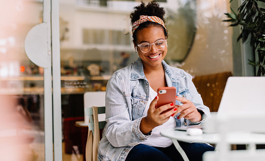 Happy woman works remotely in a café, using her smartphone for communication and social media. Her authentic smile reflects her happiness as a freelancer in the digital age.