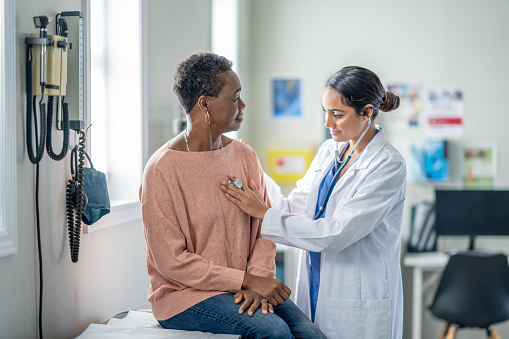 A senior woman of African decent sits up on an exam table during a routine check-up.  She is sitting still and quiet as her female doctor listens to her heart.