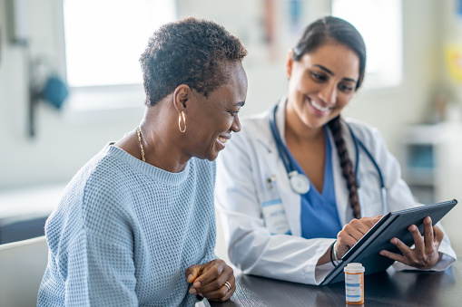 A senior woman of African decent meets with her doctor as they review her medications together.  The doctor is dressed professionally in scrubs and a lab coat and is holding out a tablet between the two as they review her recent test results together.