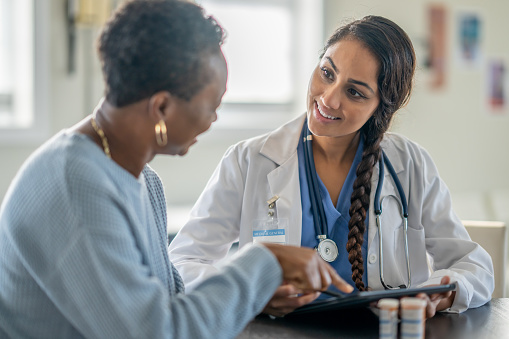 A senior woman of African decent meets with her doctor as they review her medications together.  The doctor is dressed professionally in scrubs and a lab coat and is holding out a tablet between the two as they review her recent test results together.