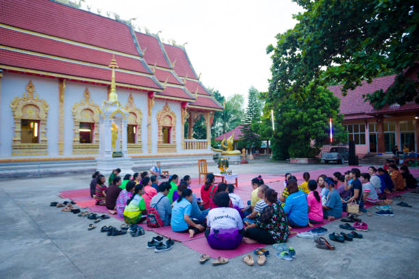 Group of adult and mature thai women at buddhist ceremony outside of a temple Group of adult and mature thai women at buddhist ceremony outside of a temple captured in Chiang Rai province in area of Chiang Khong. Women are gathering together for a local buddhist ceremony with a male musician playing classical string instrument and prayers. Women are sitting  on a plaid on floor around a buddhist flower decor. In background are people at sound mixer. The musician is sitting in front of women. At left area is temple. true thailand classic stock pictures, royalty-free photos & images