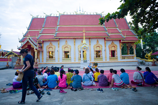 Rear view of sitting  of adult and mature thai women at buddhist ceremony outside of a temple captured in Chiang Rai province in area of Chiang Khong. Women are gathering together for a local buddhist ceremony with a male musician playing classical string instrument and prayers. Women are sitting  on a plaid on floor around a buddhist flower decor. In background is the local temple. One woman is walking in scene