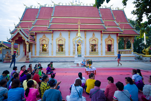 On ground sitting  thai women at buddhist ceremony outside of a temple captured in Chiang Rai province in area of Chiang Khong. Women are gathering together for a local buddhist ceremony with a male musician playing classical string instrument and prayers. Women are sitting  on a plaid on floor around a buddhist flower decor. In background is the local temple. Women are listening musician and are awaiting begin of ceremony. One woman is taking photos at temple