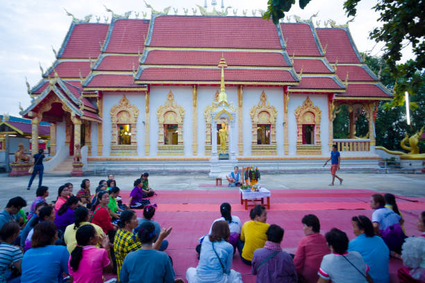 On ground sitting  thai women at buddhist ceremony outside of a temple On ground sitting  thai women at buddhist ceremony outside of a temple captured in Chiang Rai province in area of Chiang Khong. Women are gathering together for a local buddhist ceremony with a male musician playing classical string instrument and prayers. Women are sitting  on a plaid on floor around a buddhist flower decor. In background is the local temple. Women are listening musician and are awaiting begin of ceremony. One woman is taking photos at temple true thailand classic stock pictures, royalty-free photos & images