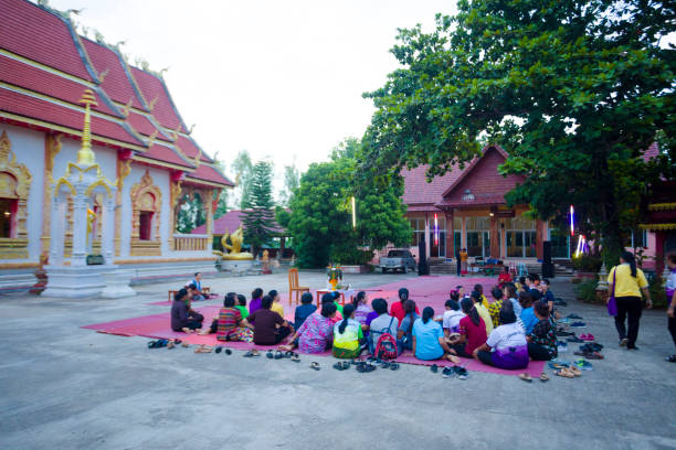 Mature thai women are listening singer women and male musician at buddhist ceremony outside of a temple Mature thai women are listening singer women and male musician at buddhist ceremony outside of a temple captured in Chiang Rai province in area of Chiang Khong. Women are gathering together for a local buddhist ceremony with a male musician playing classical string instrument and prayers. Women are sitting  on a plaid on floor around a buddhist flower decor. In background is the local temple. Temp,le is in left background true thailand classic stock pictures, royalty-free photos & images