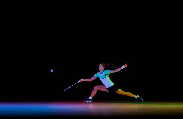 Photo of Badminton athlete demonstrates her skills in attack and defense against black background in neon light.