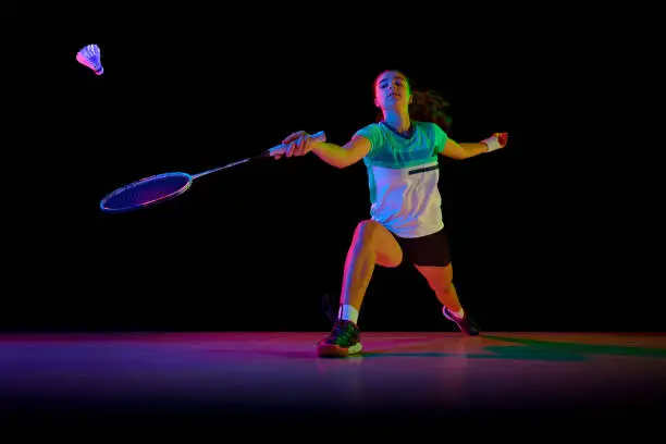 Photo of Portrait of young and attractive badminton player in motion, training diligently against black background in neon light.