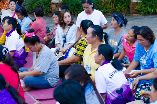 Portrait of group of mature thai women joining together at buddhist ceremony Portrait of group of mature thai women joining together at buddhist ceremony at local temple in Chiang Rai province in area of Chiang Khong. Women are gathering together for a local buddhist ceremony with a male musician playing classical string instrument and prayers. Women are sitting  on a plaid on floor around a buddhist flower decor.  Women are talking while waiting true thailand classic stock pictures, royalty-free photos & images