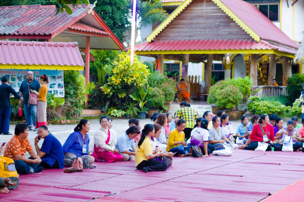 Adult and mature thai tourist women joining together at buddhist ceremony Adult and mature thai tourist women joining together at buddhist ceremony outside of  local temple in Chiang Rai province in area of Chiang Khong. Women are gathering together for a local buddhist ceremony with a male musician playing classical string instrument and prayers. Women are sitting  on a red plaids placed on floor. Women are talking while waiting or use mobile phones. In background are some more people and buildings of temple complex true thailand classic stock pictures, royalty-free photos & images