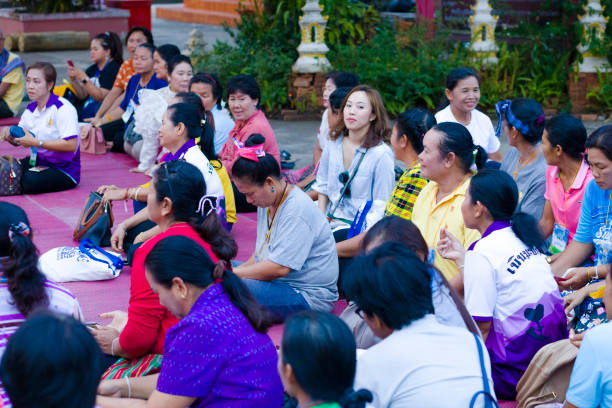 Mature thai women sitting together at buddhist ceremony Mature thai women sitting together at buddhist ceremony at local temple in Chiang Rai province in area of Chiang Khong. Women are gathering together for a local buddhist ceremony with a male musician playing classical string instrument and prayers. Women are sitting  on a plaid on floor around a buddhist flower decor.  Women are talking while waiting true thailand classic stock pictures, royalty-free photos & images