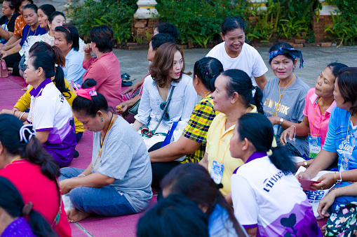 View into group of mature thai women sitting together at buddhist ceremony at local temple in Chiang Rai province in area of Chiang Khong. Women are gathering together for a local buddhist ceremony with a male musician playing classical string instrument and prayers. Women are sitting  on a plaid on floor around a buddhist flower decor.  Women are talking while waiting
