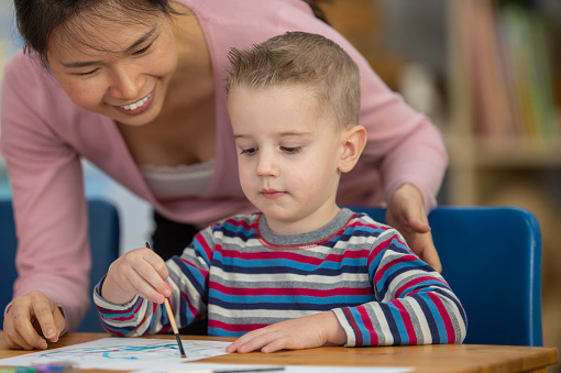 A sweet little blond haired boy sits at a desk as he focuses on painting.  His female teacher of Asian decent, is leaning in to take a closer look as his work as she encourages him in his creativity.