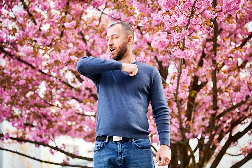 Man allergic suffering from seasonal allergy at spring in blossoming garden at springtime. Bearded young man sneezing and having runny nose in front of blooming tree. Spring allergy concept.