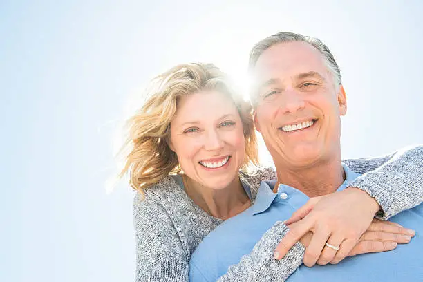 Low angle portrait of cheerful mature woman embracing man from behind against clear sky