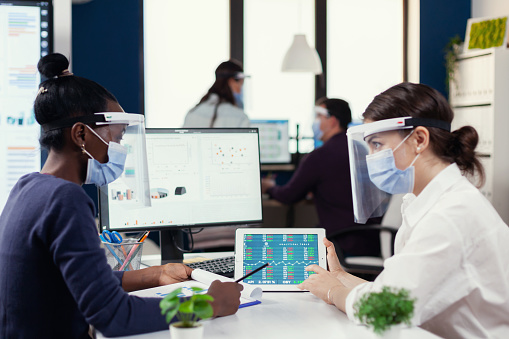 Multi ethnic employees with visor and protection mask sitting at desk analysing financial data.Diverse group of business people working and communicating together in creative office with new normal, respecting social distance.