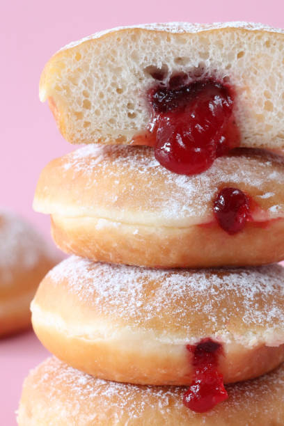 istock Full frame image of stack of fresh jam doughnuts coated in icing sugar, raspberry jam dripping from injection hole, jelly cake half, pink background, focus on foreground 1870098600