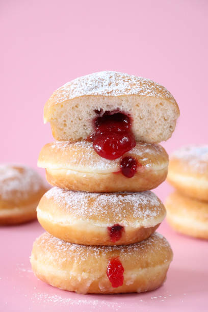 Image of stack of fresh jam doughnuts coated in icing sugar, raspberry jam dripping from injection hole, jelly cake half, pink background, focus on foreground