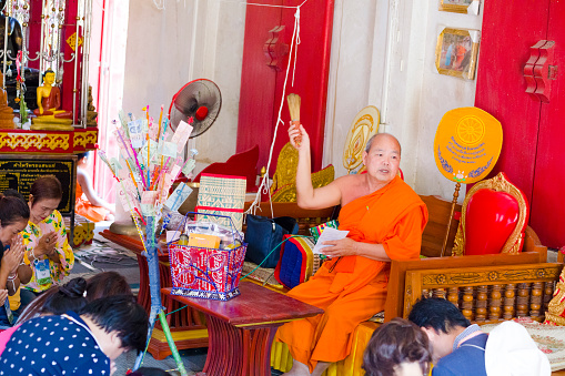 Mature monk who is sitting on a red gold colored hair is spraying water with a kind of small bamboo brush onto sitting praying people. Scene in a Buddhist ceremony with monk in thai temple  in local temple in Chiang Rai province in area of Chiang Khong..  People are mature and adult thai women who are participating at ceremony. Around monk are baskets with gifts given by women. Monk is saying a prayer