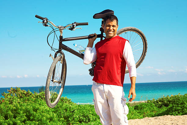 POV Point of view shot of a young latin sportsman barefoot biking a bicycle in front of a paradise blue sea landscape at Hobie Island Beach Park in Key Biscayne, Miami Beach, Miami, South Florida, United States of America - USA; wearing a blue swim shorts. On background, a beautiful sunset view over a turquoise sea and white sand.Shooting from a personal perspective in an exotic tropical beach travel holidays. Shotting location was next to Miami Seaquarium.