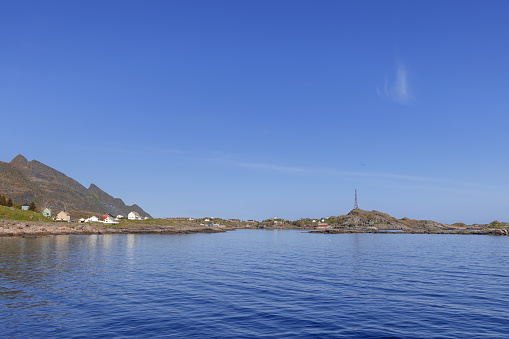 Expansive seascape showcasing traditional red Rorbu cottages along the rugged coastline of Moskenes, Lofoten Islands, Norway, bathed in summer sunshine beneath a clear azure sky