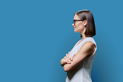 Profile view, confident successful young business woman wearing glasses with crossed arms, copy space blue studio background, space for advertising image text. Business work services education people